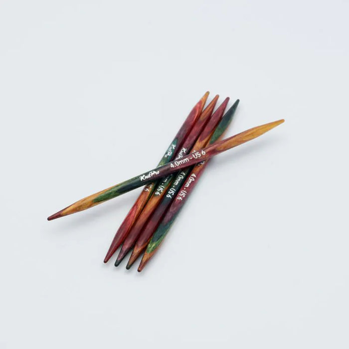 A set of six wooden knitting needles with a marbled colour pattern of red, green, and brown, crossed over each other on a light gray background. The brand name 'KnitPro' is printed on the shafts.